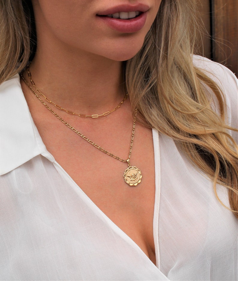 May include: A gold chain necklace with a gold pendant featuring a winged heart design. The necklace is layered over a thinner gold chain.