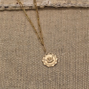 May include: A gold chain necklace with a round pendant featuring an angel with wings. The pendant has a detailed design with a scalloped edge.