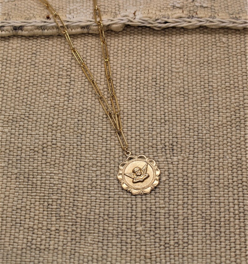 May include: A gold chain necklace with a round pendant featuring an angel with wings. The pendant has a detailed design with a scalloped edge.