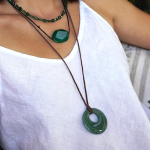 May include: A woman wearing three green gemstone necklaces. The top necklace is a delicate strand of small beads. The middle necklace features a large, faceted oval pendant. The bottom necklace has a large, circular pendant with a hole in the center.