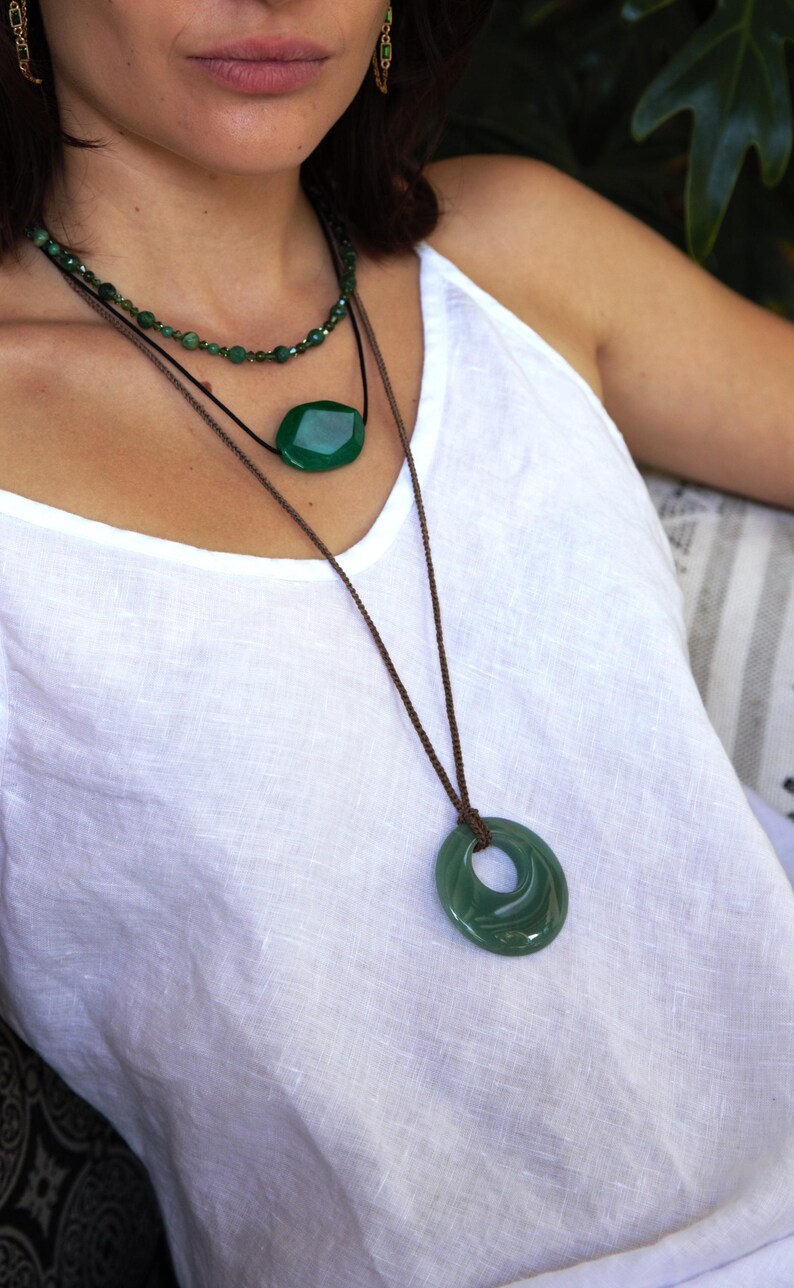 May include: A woman wearing three green gemstone necklaces. The top necklace is a delicate strand of small beads. The middle necklace features a large, faceted oval pendant. The bottom necklace has a large, circular pendant with a hole in the center.