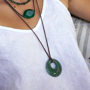 Op de afbeelding: Een gelaagde ketting set met drie strengen groene edelsteen hangers. De bovenste streng heeft kleine kralen, de middelste heeft een grote ovale steen en de onderste heeft een grote, open cirkel hanger, allemaal op bruine koorden.