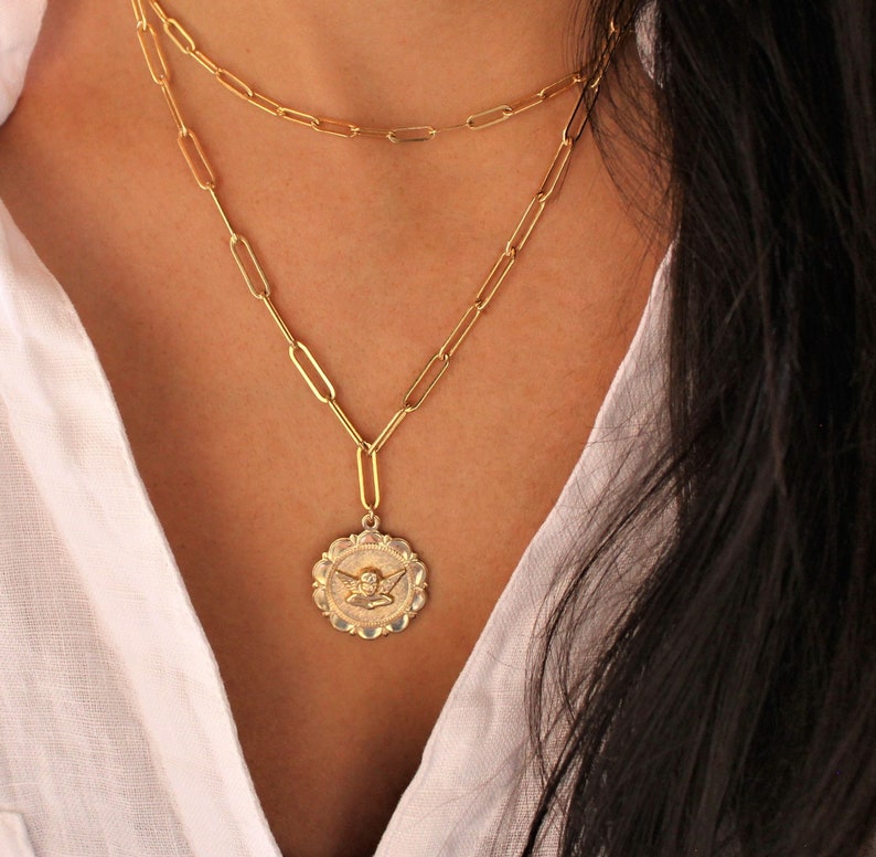 May include: A gold chain necklace with a gold angel pendant. The chain is made of linked ovals and the pendant is round with a detailed angel design.
