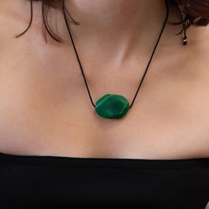 May include: A green gemstone pendant necklace. The pendant is a faceted, irregular shape, suspended from a black cord. The necklace is worn against a person's skin, with a black strapless top visible.