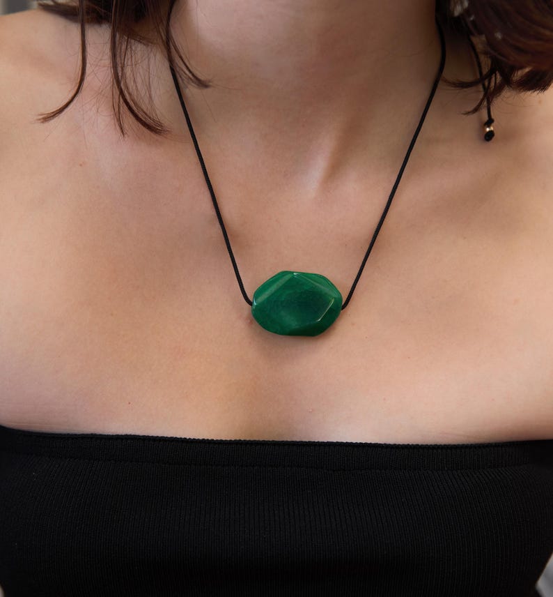 May include: A green gemstone pendant necklace. The pendant is a faceted, irregular shape, suspended from a black cord. The necklace is worn against a person's skin, with a black strapless top visible.
