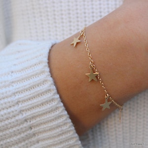 May include: A gold chain bracelet with dangling star charms. The bracelet is on a person's wrist.