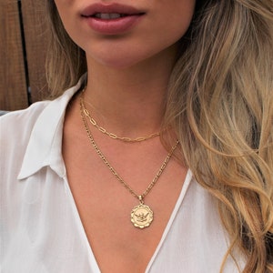 May include: A gold chain necklace with a pendant featuring a detailed angel design. The necklace is layered with a second, thinner chain.