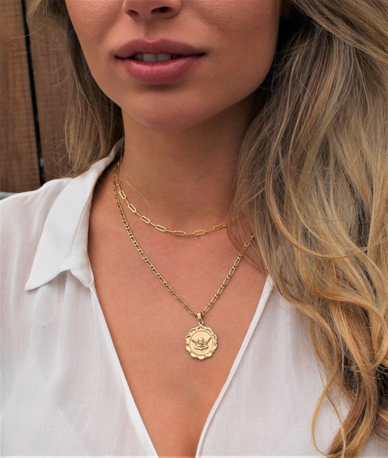 May include: A gold chain necklace with a pendant featuring a detailed angel design. The necklace is layered with a second, thinner chain.