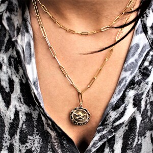 May include: A gold chain necklace with a pendant featuring a detailed design. The necklace is layered with two other gold chains.