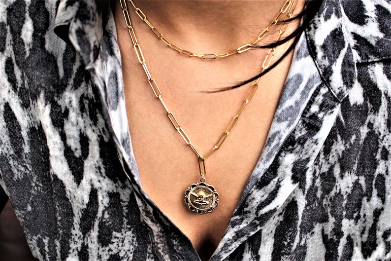 May include: A gold chain necklace with a pendant featuring a detailed design. The necklace is layered with two other gold chains.