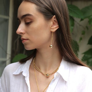 May include: A woman wearing a white shirt with a gold layered necklace and matching gold earrings with a round pendant.