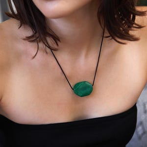 May include: A green gemstone pendant necklace. The pendant is an irregularly shaped, faceted stone suspended from a black cord. The necklace is worn against a black top.