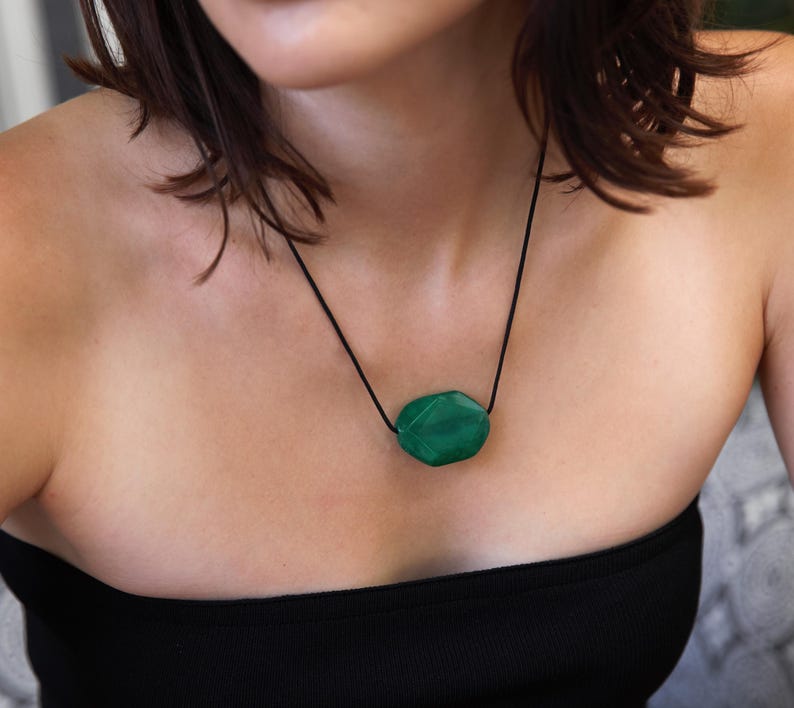 May include: A green gemstone pendant necklace. The pendant is an irregularly shaped, faceted stone suspended from a black cord. The necklace is worn against a black top.