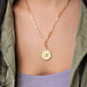 May include: A gold chain necklace with a gold pendant featuring an angel design. The necklace is worn by a person with long dark hair and a green jacket.