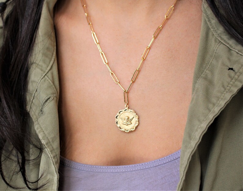 May include: A gold chain necklace with a gold pendant featuring an angel design. The necklace is worn by a person with long dark hair and a green jacket.