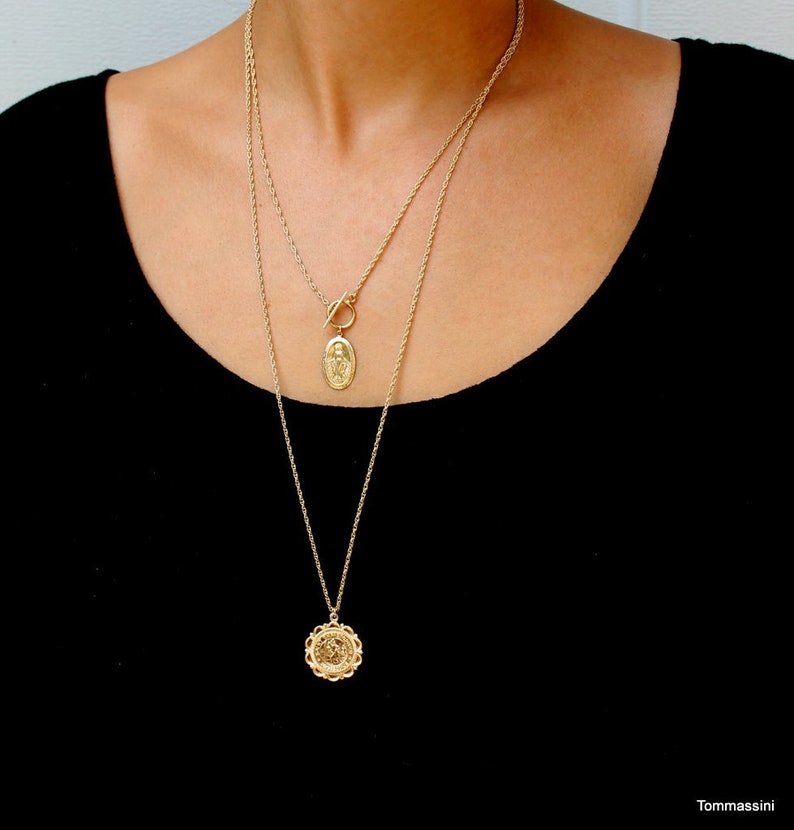 May include: Two gold chain necklaces with pendants. The top necklace has a toggle clasp and a small oval pendant with a religious figure. The bottom necklace has a larger round pendant with a detailed design.