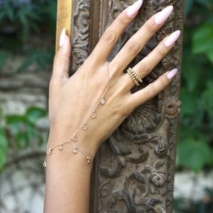 Könnte beinhalten: Eine zarte goldene Handkette mit kleinen, funkelnden, klaren Steinen hängt vom Handgelenk und über dem Handrücken. Ein goldener Ring mit drei Reihen kleiner Perlen befindet sich am Ringfinger. Die Nägel sind hellrosa lackiert.