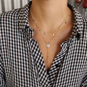 Puede incluir: Un collar de cadena de oro con un peque&ntilde;o colgante de cruz y un colgante m&aacute;s grande con un icono religioso. El collar est&aacute; superpuesto con una segunda cadena con peque&ntilde;os colgantes de cruz.