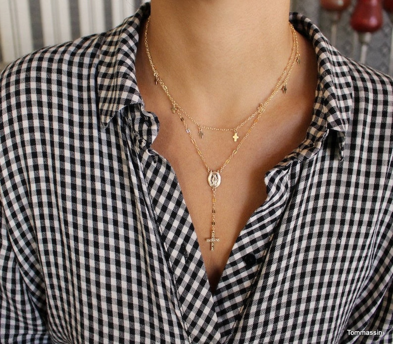 Puede incluir: Un collar de cadena de oro con un peque&ntilde;o colgante de cruz y un colgante m&aacute;s grande con un icono religioso. El collar est&aacute; superpuesto con una segunda cadena con peque&ntilde;os colgantes de cruz.