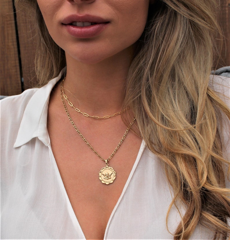 May include: A gold chain necklace with a delicate pendant featuring a stylized angel design. The necklace is layered with a second, thinner chain.