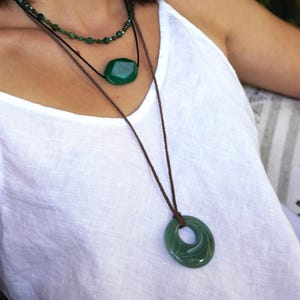 May include: A woman wearing three green gemstone necklaces. The top necklace is a beaded strand, the middle one features a large, faceted stone, and the bottom one has a circular pendant. The necklaces are layered and hang against a white top.