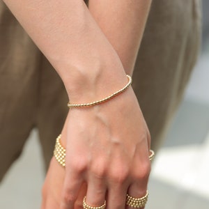 May include: A gold beaded bracelet is worn on a wrist, with other gold beaded jewelry visible. The bracelet is made of small, round beads strung together. The jewelry is a warm, shiny gold color.