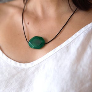 May include: A faceted, emerald green gemstone pendant necklace. The large, irregularly shaped stone is suspended from a thin, black cord. The necklace is displayed against a white background.