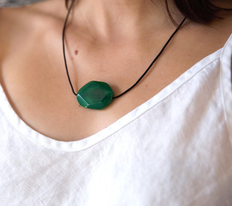 May include: A faceted, emerald green gemstone pendant necklace. The large, irregularly shaped stone is suspended from a thin, black cord. The necklace is displayed against a white background.