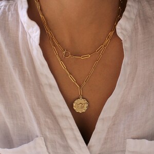 May include: A gold chain necklace with a gold pendant featuring an angel design. The necklace is layered with a second chain with a toggle clasp.