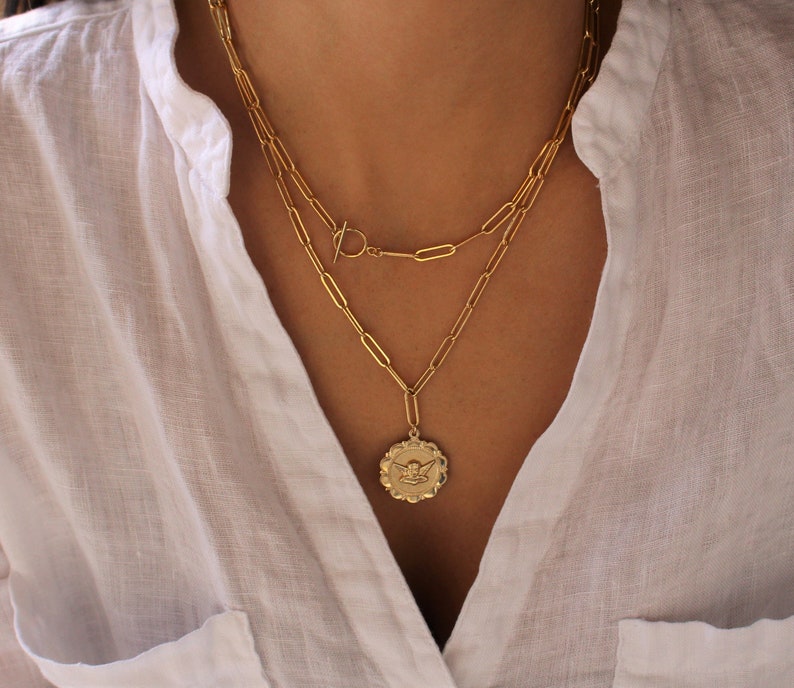 May include: A gold chain necklace with a gold pendant featuring an angel design. The necklace is layered with a second chain with a toggle clasp.
