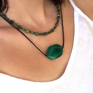 May include: A layered necklace featuring two strands. The top strand is composed of small, green beads. The lower strand showcases a large, faceted, emerald-green gemstone pendant suspended from a black cord. The necklace is worn against a white top.
