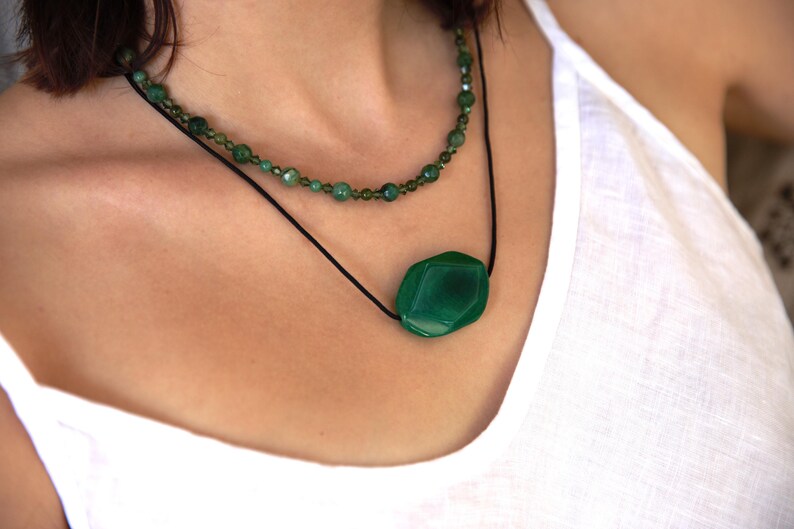 May include: A layered necklace featuring two strands. The top strand is composed of small, green beads. The lower strand showcases a large, faceted, emerald-green gemstone pendant suspended from a black cord. The necklace is worn against a white top.