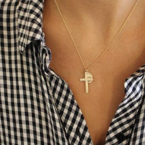 Puede incluir: Un collar de cadena dorada con un colgante de cruz y un pequeño dije redondo. El collar lo lleva una persona que lleva una camisa a cuadros blancos y negros.