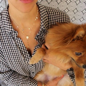 Puede incluir: Una mujer con una camisa de cuadros blancos y negros est&aacute; sosteniendo un perro peque&ntilde;o, esponjoso y marr&oacute;n claro. Lleva un collar de oro con un peque&ntilde;o colgante de cruz y una cadena de capas con abalorios de estrellas.