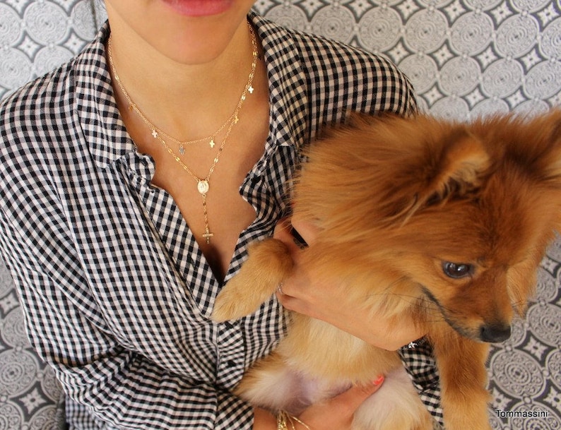 Puede incluir: Una mujer con una camisa de cuadros blancos y negros est&aacute; sosteniendo un perro peque&ntilde;o, esponjoso y marr&oacute;n claro. Lleva un collar de oro con un peque&ntilde;o colgante de cruz y una cadena de capas con abalorios de estrellas.