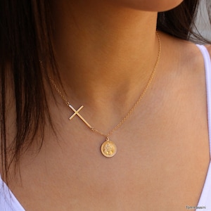 Puede incluir: Un collar de cadena dorada con un colgante de cruz lateral y un colgante de moneda redonda.