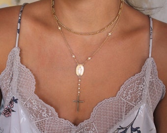May include: A gold chain necklace with a small oval pendant featuring a religious image and a cross charm. The necklace is layered with a second gold chain.