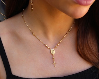 May include: A gold-toned rosary necklace featuring a delicate chain with small round beads and elongated bar links. The necklace includes a central oval pendant depicting a figure and a small cross charm hanging at the bottom.