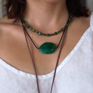 May include: A layered necklace set featuring three strands. The top strand is a beaded necklace with small green beads. The middle strand has a large, oval-shaped green stone pendant. The bottom strand is a long, brown chain.