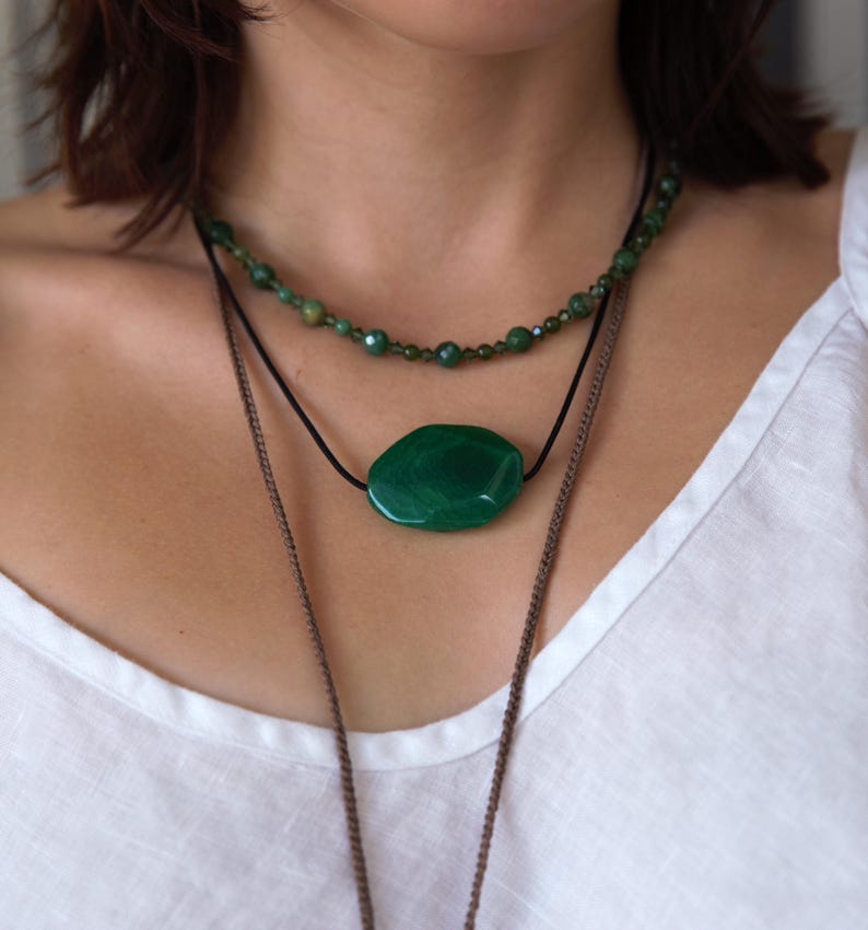 May include: A layered necklace set featuring three strands. The top strand is a beaded necklace with small green beads. The middle strand has a large, oval-shaped green stone pendant. The bottom strand is a long, brown chain.