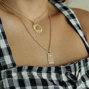 May include: Two gold necklaces with pendants. The top necklace has a round pendant with a detailed design. The bottom necklace has a rectangular pendant with an inscription.