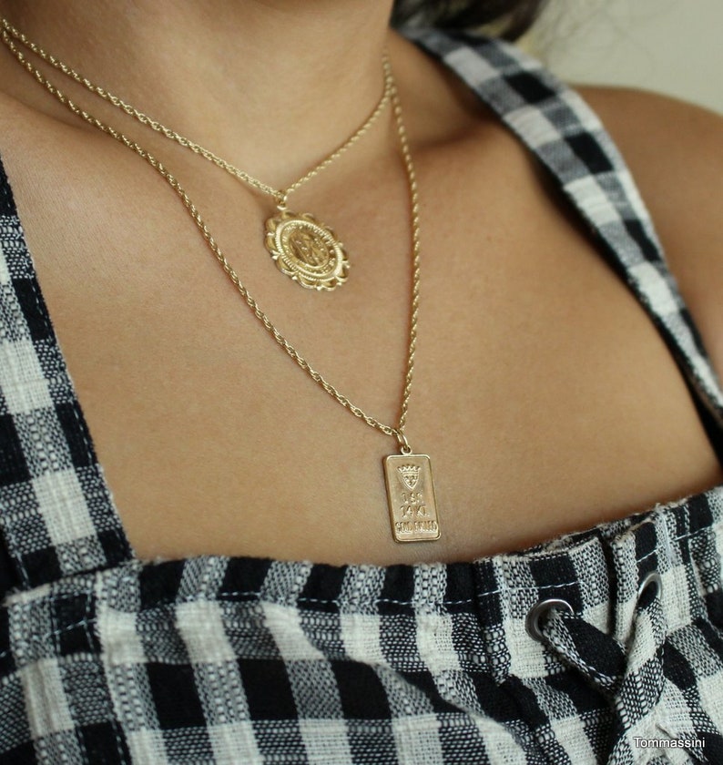 May include: Two gold necklaces with pendants. The top necklace has a round pendant with a detailed design. The bottom necklace has a rectangular pendant with an inscription.