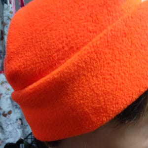 Könnte beinhalten: Eine leuchtend orangefarbene Fleece-Mütze. Die Mütze ist aus weichem, flauschigem Material und hat ein schlichtes Design.