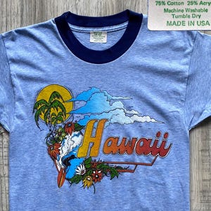 Puede incluir: Una camiseta ringer azul claro con ribetes azul oscuro en el cuello y las mangas. El frente presenta un gráfico de palmeras, flores y nubes con el texto Hawaii. Una etiqueta indica: MEDIUM (38-40), 75% Cotton 25% Acrylic, Machine Washable, Tumble Dry, MADE IN USA.