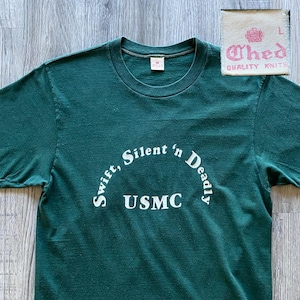 May include: A dark green t-shirt with the text "Swift, Silent 'n Deadly" arched above "USMC" in white. The shirt has short sleeves and a crew neck. A small tag with the text "Ched Quality Knits" is in the upper right corner.
