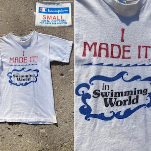 Könnte beinhalten: Weißes T-Shirt mit einer roten und blauen Grafik, auf der "I MADE IT! in Swimming World" steht. Das Shirt ist von der Marke Champion und hat die Größe Small.