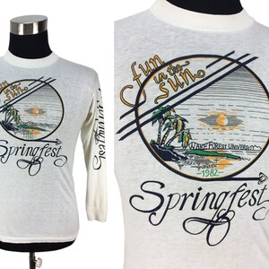 May include: White long-sleeve t-shirt with a graphic of a palm tree scene in a circle with the text "Fun in the Sun" and "Springfest '82" underneath. The text "Wake Forest University" is also included in the graphic.
