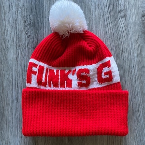 May include: A red knit beanie with a white band featuring the text "FUNK'S G" in bold white letters. The hat has a white pom-pom on top.