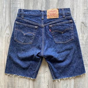 Könnte beinhalten: Dunkelblaue Jeansshorts mit rohem Saum. Die Shorts verfügen über ein klassisches Fünf-Taschen-Design, einschließlich zweier Gesäßtaschen mit der charakteristischen Levi's-Bogennaht. Ein rotes Levi's-Etikett ist auf der Gesäßtasche sichtbar.