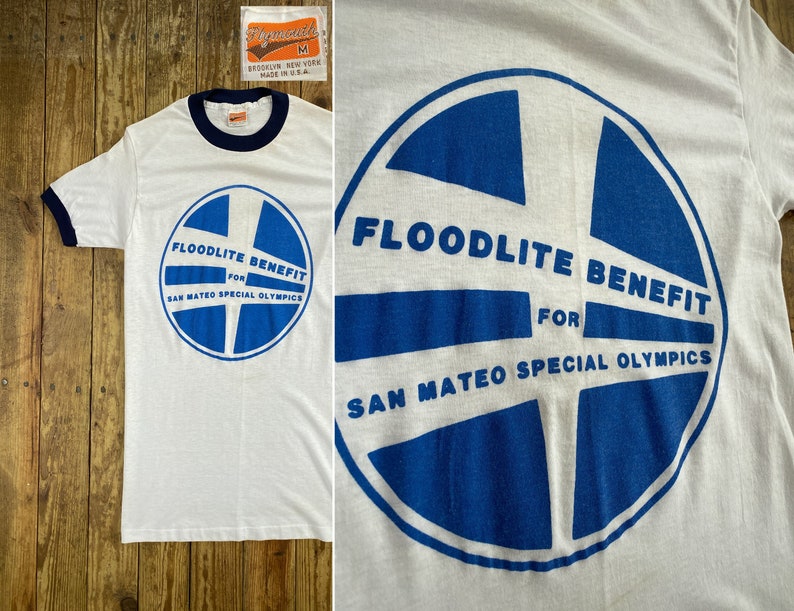 Pu&ograve; includere: Maglietta bianca con un grafico circolare blu e bianco che dice "Floodlite Benefit for San Mateo Special Olympics".